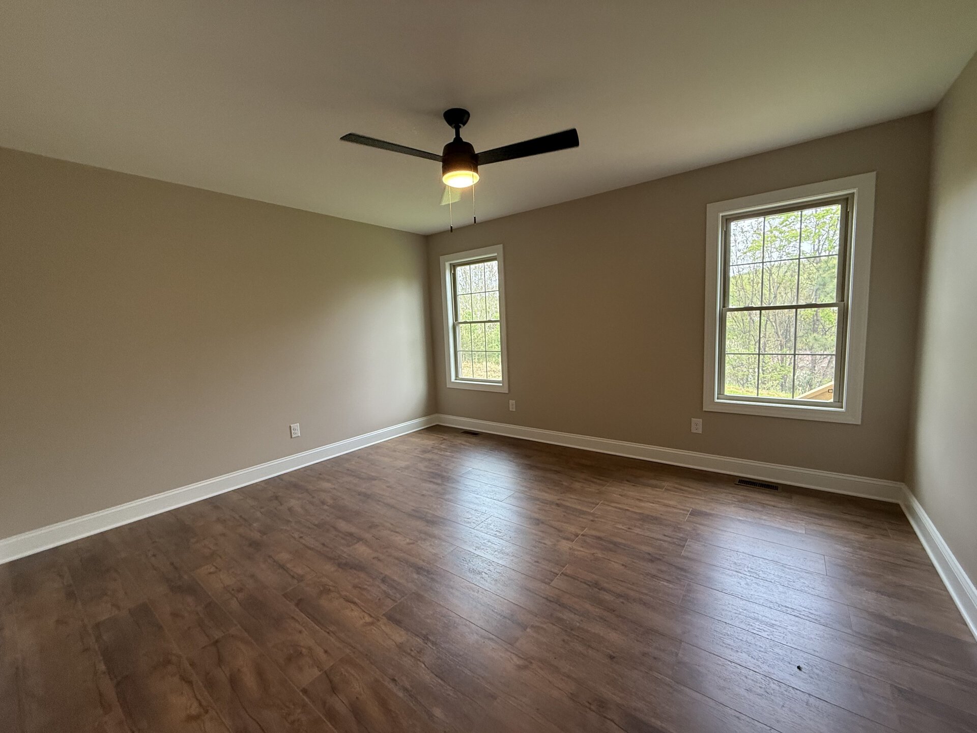 Bright empty room with two windows, ceiling fan, and hardwood flooring.