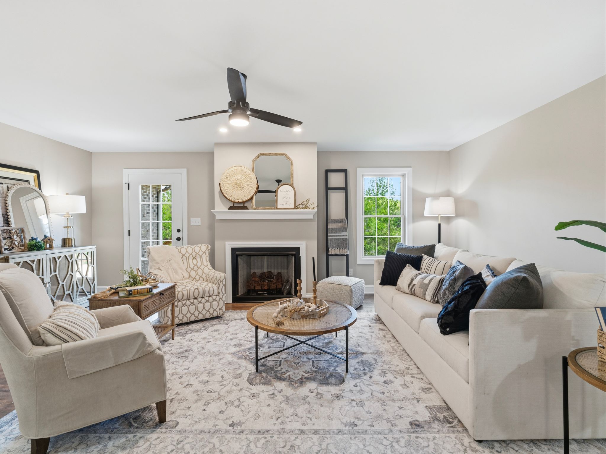 Bright living room with fireplace, sofas, and large windows showing greenery.