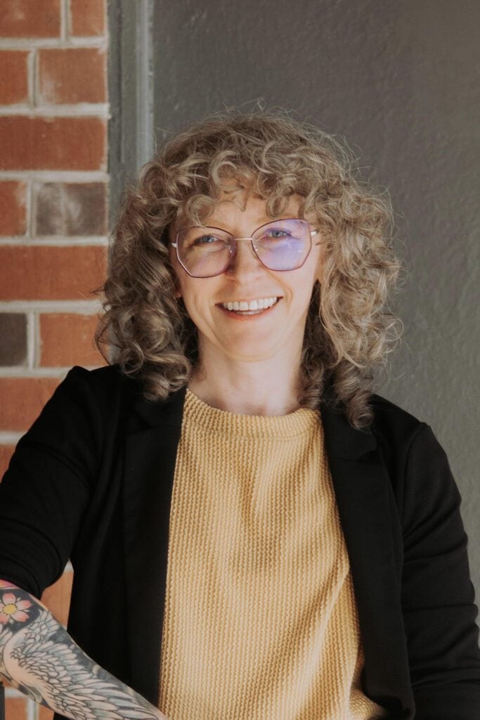 Smiling woman with curly hair and glasses in front of a brick wall.